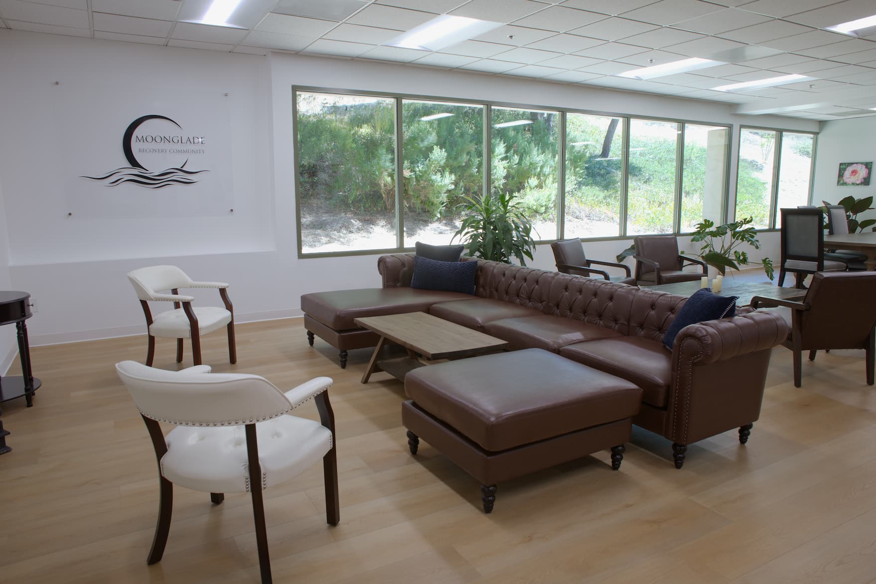 Window-lined Moonglade Recovery common room with leather sofas, armchairs, and views of greenery